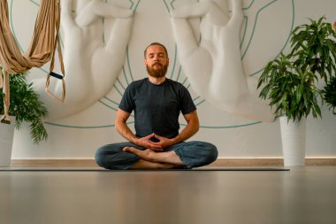 Sakallı yetişkin bir adam ellerini önünde tutuyor ve gözleri kapalı hafif yoga stüdyosundaki lotus pozisyonunda meditasyon yapıyor.