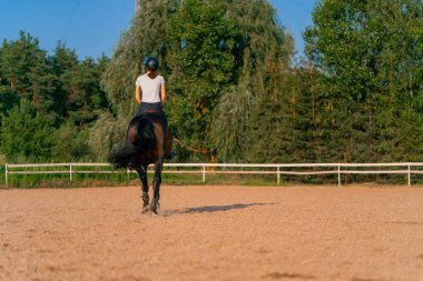 Miğfer takmış bir atlı kadın ata binerken arenada güzel siyah atına biniyor.
