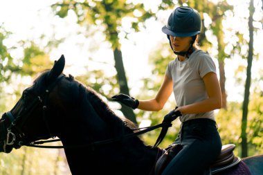 Miğfer takmış bir binici at sürerken ormanda güzel siyah atına biner.