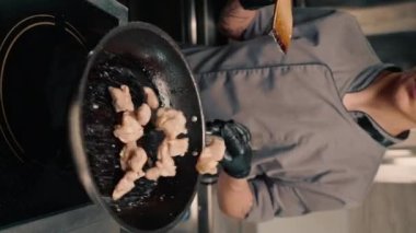 Bir restoranda, profesyonel bir mutfakta şefin tavaya yakın dikey bir videoda sulu tavuk filetosu pişirmesi.