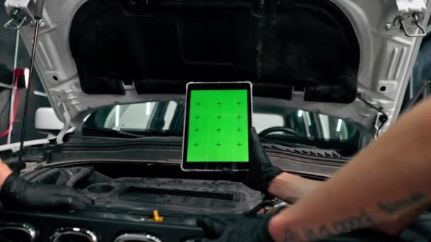 フードが開いている車の背景に対して緑のスクリーンが開いているタブレットが付いている手のクローズアップ — ストック動画