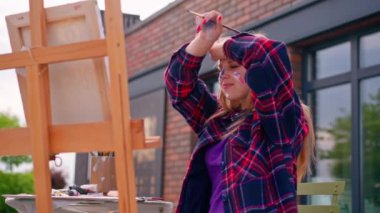 Yüzünde boya olan odaklanmış bir kız ressam boya fırçasıyla sehpada duran bir resim çiziyor.