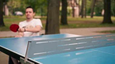 Tekerlekli sandalyedeki bir adam şehir parkında masa tenisi oynuyor pinpon raketiyle vuruşlar yapıyor.