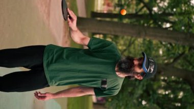 dikey video yetişkin sakallı adam şehir parkında pinpon oynadıktan sonra tenis raketiyle turuncu bir topa vuruyor.