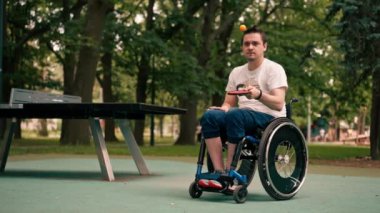 Tekerlekli sandalyedeki bir adam, şehir parkında pinpon oynadıktan sonra tenis raketiyle turuncu bir topa vurmaya konsantre olur.