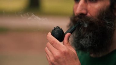 Yakın plan sakallı bir adam, elinde tütünle şehir parkında sigara tüttürüyor, sigara içiyor, kötü alışkanlıklar ediniyor.
