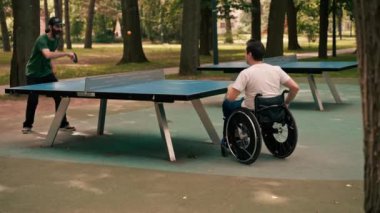 Tekerlekli sandalyedeki bir adam şehir parkında arkadaşıyla masa tenisi oynuyor.