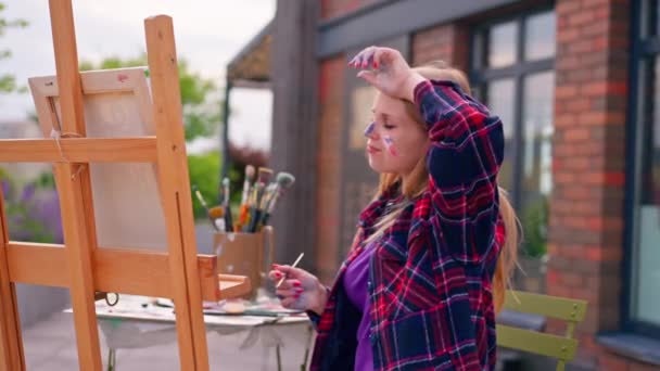 ペイントブラシでイーゼルに立っている絵を描く彼女の顔にペンキをかけた少女アーティスト — ストック動画
