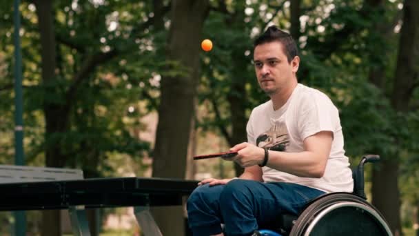 車椅子の男は 都市公園でピンポンをプレイした後 テニスラケットでオレンジ色のボールを打つことに集中します — ストック動画