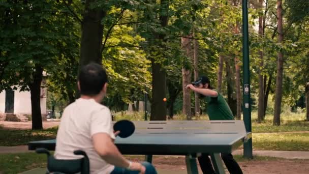 車椅子の男は 都市公園の野外活動で彼の友人と卓球ピンポンを果たしています — ストック動画