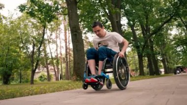 Gülümseyen genç adam, şehir parkında aktif bir yaşam tarzında tekerlekli sandalyede numaralar yapıyor.