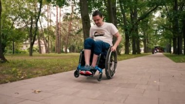 Gülümseyen genç adam, şehir parkında aktif bir yaşam tarzında tekerlekli sandalyede numaralar yapıyor.