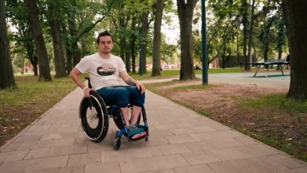 車椅子の若い男が市立公園を歩き 路地に立ち寄り 意図的にカメラを見ている — ストック動画