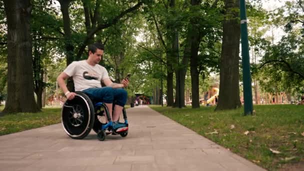 車椅子の若い男は 感情的にニュースを読み取ったり 都市公園のテクノロジーモビリティのメッセンジャーで通信したりする意図的に電話を見ます — ストック動画