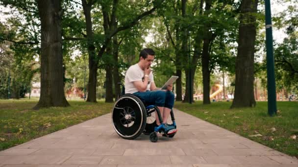 坐在轮椅上的年轻人聚精会神地看着平板电脑 若有所思地阅读城市公园技术流动的新闻 — 图库视频影像