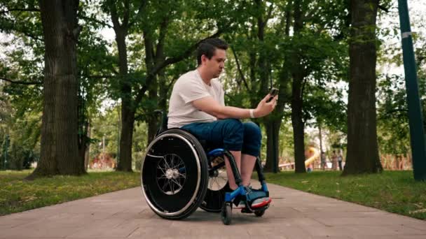 坐在轮椅上的年轻人聚精会神地看着电话 感情用事地阅读新闻或在城市公园技术流动的信使中进行交流 — 图库视频影像