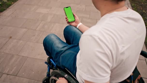 Man Rullstol Håller Telefon Med Öppen Grön Skärm Stadsparken Bakifrån — Stockvideo