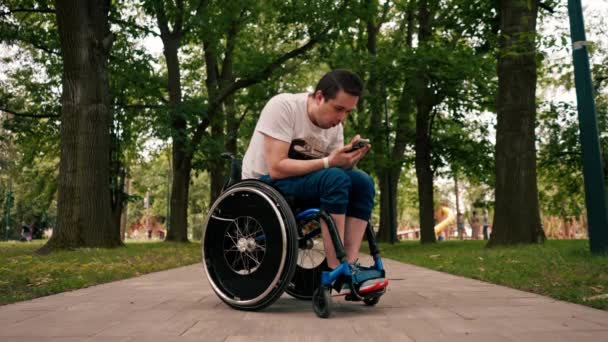 坐在轮椅上的年轻人聚精会神地看着电话 感情用事地阅读新闻或在城市公园技术流动的信使中进行交流 — 图库视频影像