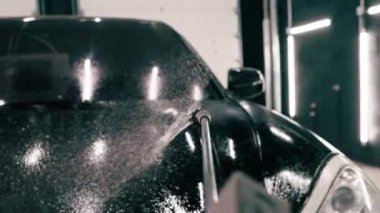 Erkek oto yıkama çalışanı oto yıkama kutusunda dikey olarak sprey tabanca kullanan siyah lüks bir arabaya araba deterjanı uyguluyor.