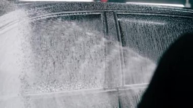 Erkek oto yıkama çalışanı oto yıkama kutusundaki sprey tabancayı kullanarak siyah lüks bir arabaya araba yıkama deterjanı sürüyor.