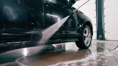 Erkek oto yıkama çalışanı oto yıkama kutusundaki sprey tabancayla siyah lüks bir arabaya deterjan uyguluyor.