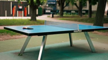İki tenis raketi ve turuncu bir tenis topu, mavi bir tenis masasının üzerinde şehir parkındaki masa tenisi oyununun ağlarının yanında yatıyor.