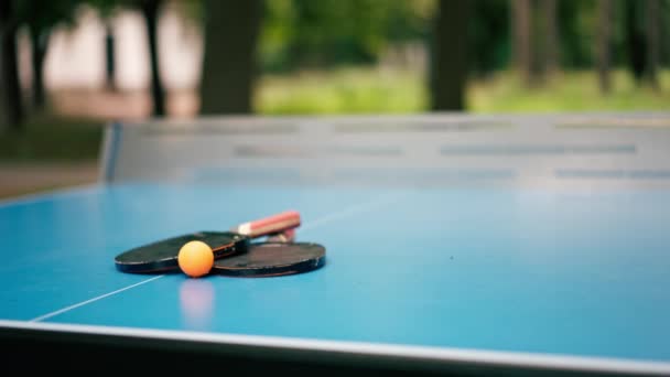 Две Теннисные Ракетки Оранжевый Теннисный Мяч Лежат Синем Теннисном Столе — стоковое видео