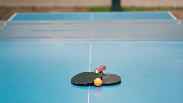 在一个城市公园的乒乓球比赛中 两个网球拍和一个橙色的网球球躺在一张蓝色的网球桌上 — 图库视频影像