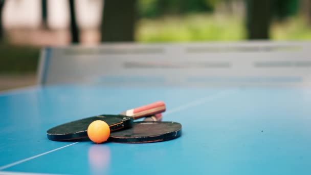 クローズアップ つのテニスラケットとオレンジテニスボールは 都市公園のピンポンゲームのネットの隣にある青いテニステーブルに横たわっています — ストック動画