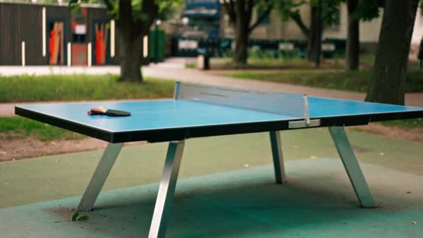 2つのテニスラケットとオレンジテニスボールは 都市公園のピンポンゲームのネットの隣にある青いテニステーブルの上にあります — ストック動画
