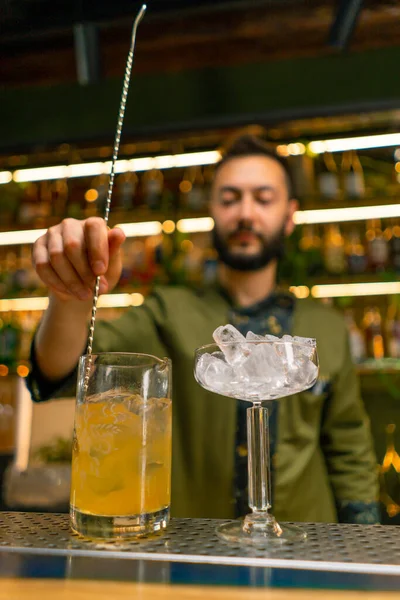 A classic bartender stirs alcohol in a tall glass with a long spoon to prepare cocktail at the bar