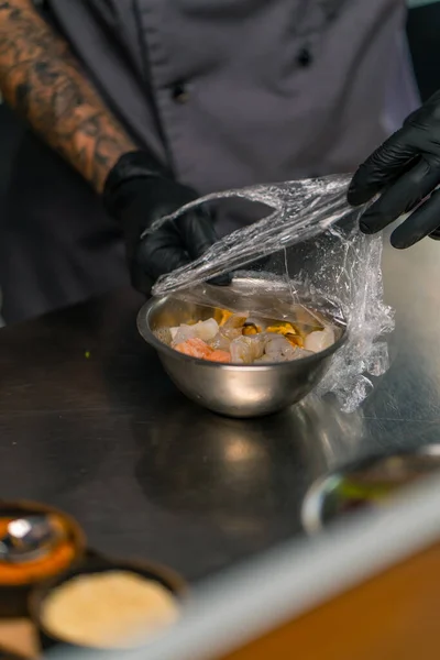 close-up of a chef in black gloves removing cling film from plate containing seafood before cooking