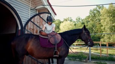 Profesyonel kıyafetli bir kadın binici, ormanda yürüyüş yapmadan önce ahırın yakınındaki güzel atına biner. Hayvanları sever, binicilik sporu yapar.