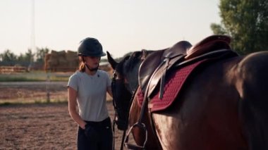 Miğferli bir sürücü siyah atını okşuyor. Yürüyüş sırasında at binme çiftliğindeki bir arenada ekipmanları düzeltiyor.