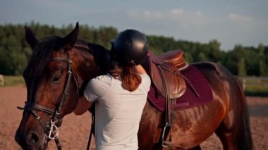 Miğferli bir sürücü siyah atını okşuyor. Yürüyüş sırasında at binme çiftliğindeki bir arenada ekipmanları düzeltiyor.