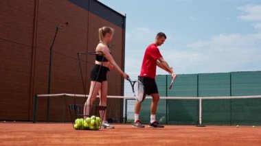 Genç bir koç, acemi bir kıza tenis oynamayı öğretir, tenis kortunda tenis topuyla raket pratiği yapar.