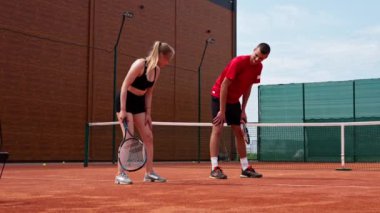 Genç bir koç, acemi bir kıza tenis oynamayı öğretir, tenis kortunda tenis topuyla raket pratiği yapar.