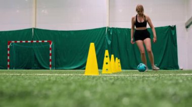 Genç kız futbol sahasında antrenman yapıyor topu cipslerin ya da konilerin arasına sürüyor ve maça hazırlanıyor.