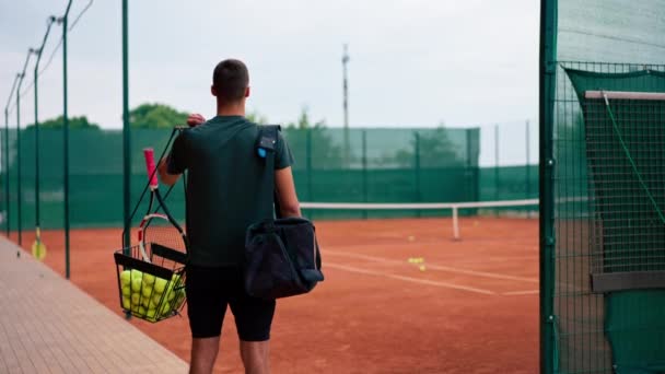 ラケットとバスケットテニスボールのバックビューでゲームトレーニングを開始する前に屋外テニスコートに立っている若いプロの選手コーチ — ストック動画
