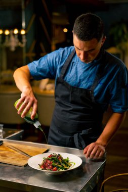 İtalyan restoranındaki üniformalı şef elinde bir şişe sos tutuyor ve mutfakta hazırladığı salatayı giydiriyor.