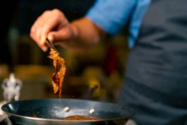 İtalyan restoranının profesyonel mutfağında elinde maşayla domuz pastırması kızartan bir şefin yakın çekimi.
