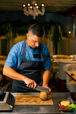 Bir şef, restoranda profesyonel bir mutfakta hamburger pişirirken ekmeği bıçakla keser.
