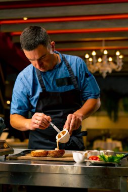 Profesyonel bir restoran mutfağında hamburger pişirirken çöreğin üzerine sos döken şefin yakın çekimi.