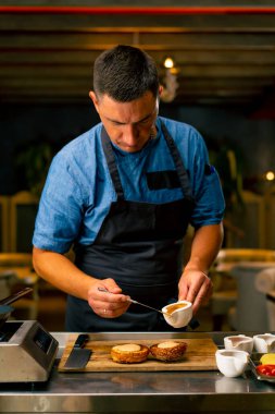 Profesyonel bir restoran mutfağında hamburger pişirirken çöreğin üzerine sos döken şefin yakın çekimi.