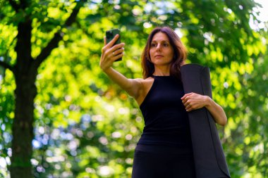 Eğitimden önce ya da sonra parkta yürüyüş yapan aktif, güzel bir yogi kadın telefonda fotoğraf çekerken elinde spor paspası tutuyor.