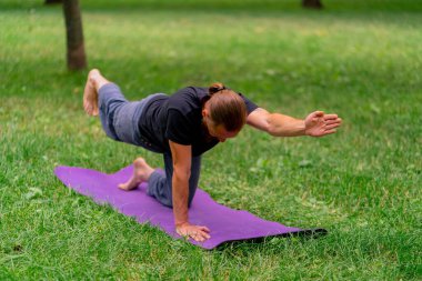 Yakışıklı sakallı adam parkta meditasyon yapıyor yoga yapıyor esneme hareketleri yapıyor ve farklı kas grupları çalıştırıyor.