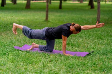 Yakışıklı sakallı adam parkta meditasyon yapıyor yoga yapıyor esneme hareketleri yapıyor ve farklı kas grupları çalıştırıyor.