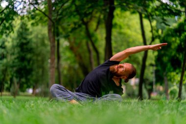 Yakışıklı sakallı adam parkta meditasyon yapıyor yoga yapıyor esneme hareketleri yapıyor ve farklı kas grupları çalıştırıyor.