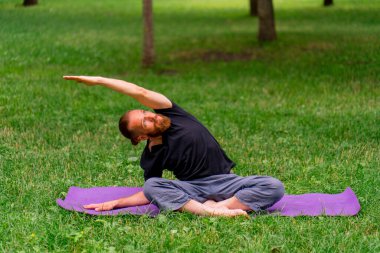 Yakışıklı sakallı adam parkta meditasyon yapıyor yoga yapıyor esneme hareketleri yapıyor ve farklı kas grupları çalıştırıyor.