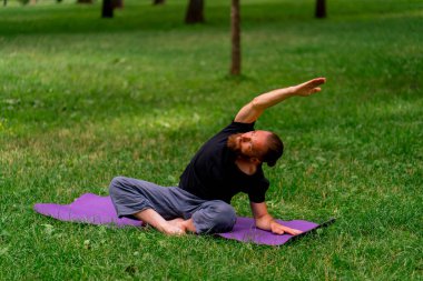 Yakışıklı sakallı adam parkta meditasyon yapıyor yoga yapıyor esneme hareketleri yapıyor ve farklı kas grupları çalıştırıyor.